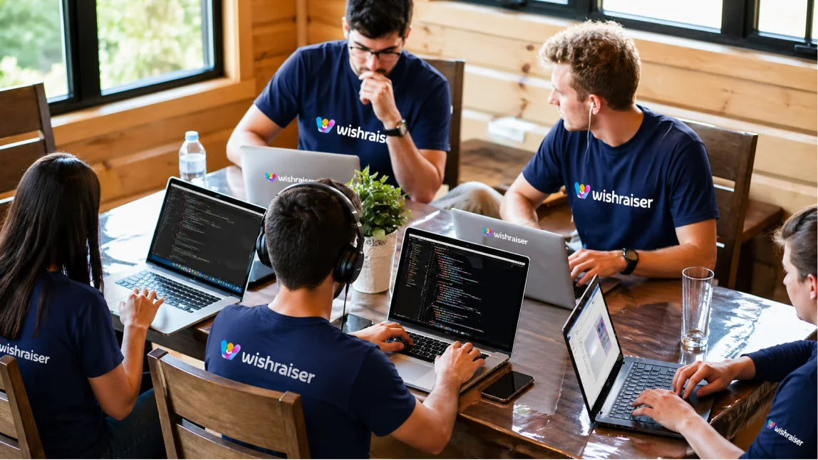 Gruppo di persone sedute attorno a un tavolo mentre lavorano con laptop in un ufficio in legno; indossano magliette blu con logo Wishraiser e sugli schermi è visibile codice di programmazione.